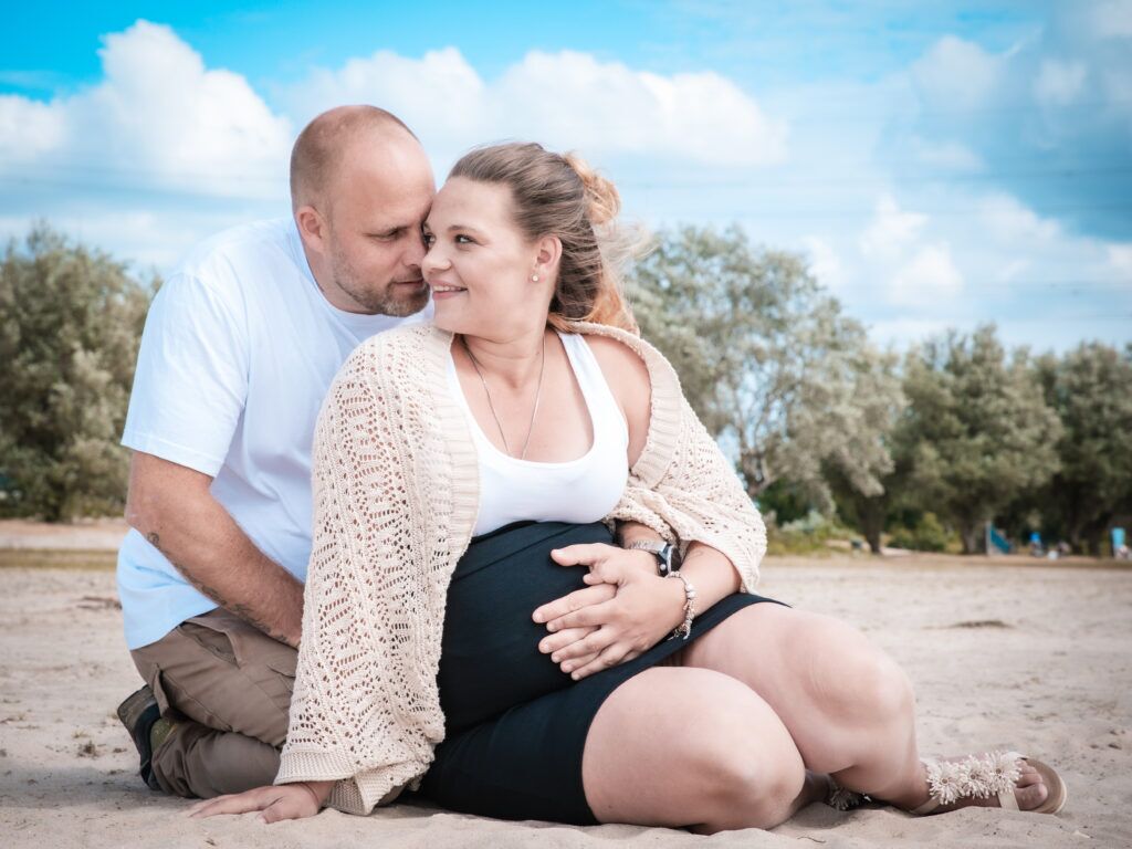 Koppel is zwanger en kijkt mekaar liefdevol aan hurkend op het strand