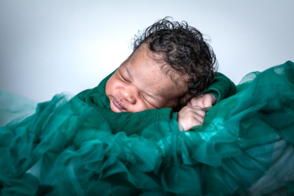 Een baby slaapt vredig op een zacht kussen, bedekt door de vloeiende jurk van de moeder. De scène straalt warmte, geborgenheid en rust uit, met een dromerige en serene sfeer.