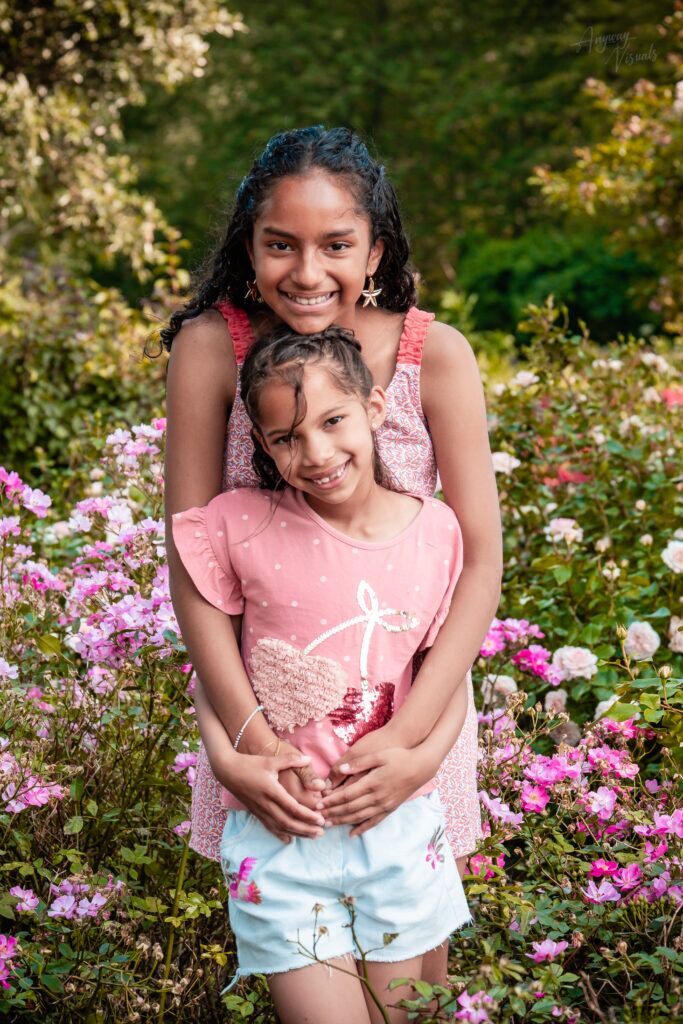 Twee zusjes staan achter elkaar en omhelzen elkaar liefdevol, met een natuurlijke omgeving en bloemen op de achtergrond