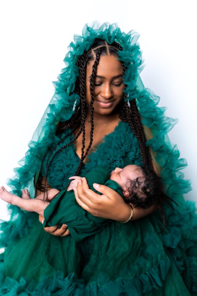 Een liefdevolle moeder met gesloten ogen knuffelt haar baby teder, terwijl ze het iets lager in haar armen houdt. Ze draagt een elegante jurk en straalt warmte en geborgenheid uit tegen een zachte, dromerige achtergrond.